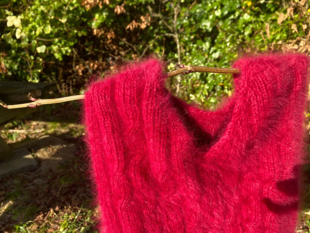 Een slipover (in wording) van rode angorawol, gefotografeerd in de tuin met een takje als kleerhanger..